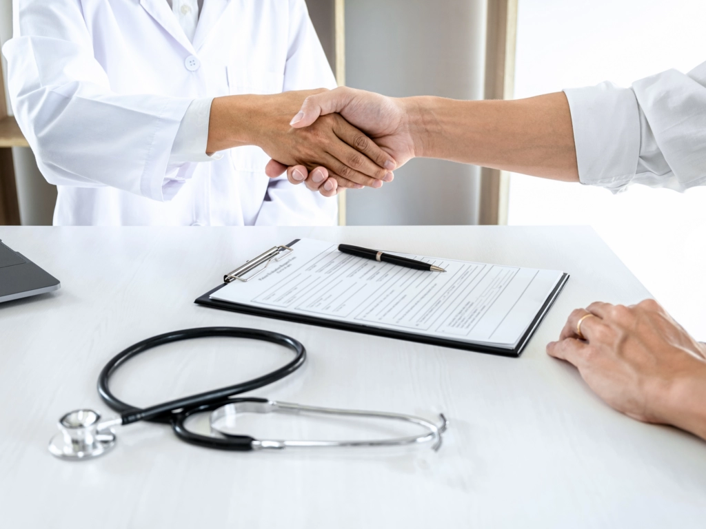 Arzt und Patient geben sich über einem Schreibtisch mit Formularen und Stethoskop die Hand.