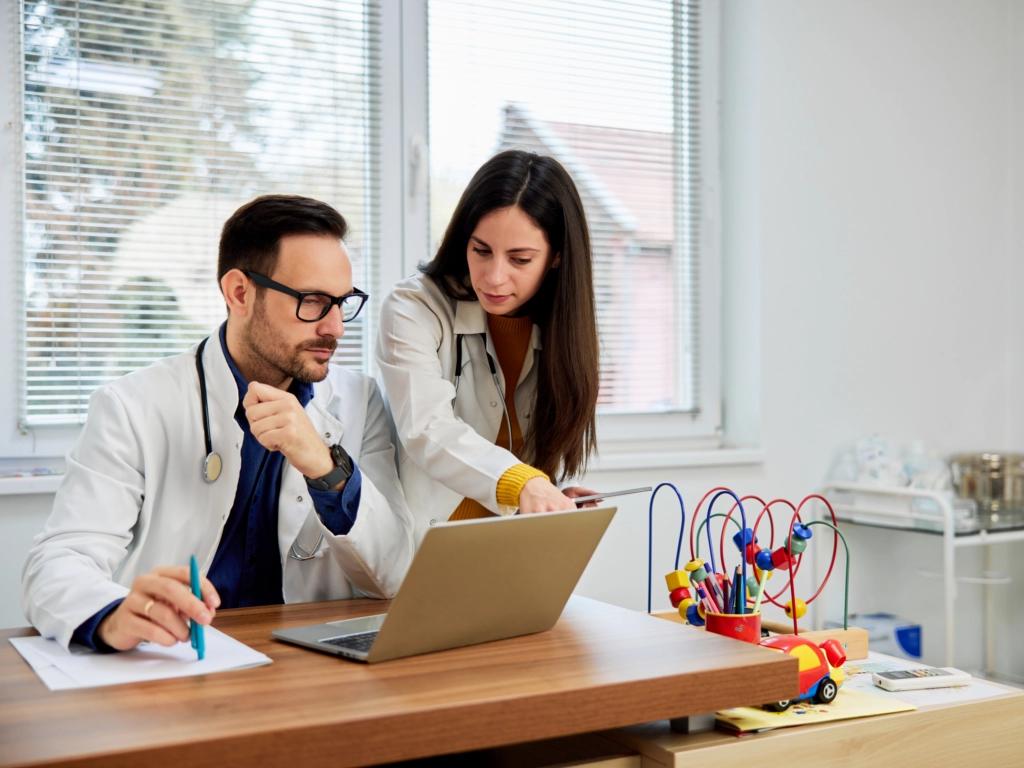 Arzt und Kollegin besprechen gemeinsam Informationen auf einem Laptop in einem hellen Behandlungszimmer.